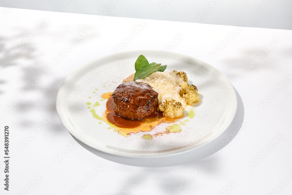 Beef cutlet with grilled cauliflower with gravy and cheese sauce on white plate. Elegant beefsteak on light background with shadows of sunlight. Summer menu with hard shadows Meat ball of minced meat