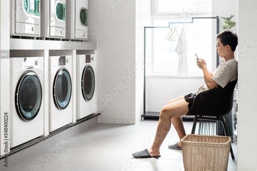 Slika na platnu Asian young man is sitting in the public laundry room, using mobile phone while waiting for the laundry process