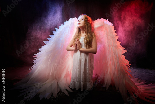 A girl in a white shirt, wings and with long hair, looking like a angel. A young model posing at a photo shoot with smoke, fog, flour, dust. A little fairy girl on a dark black background photo