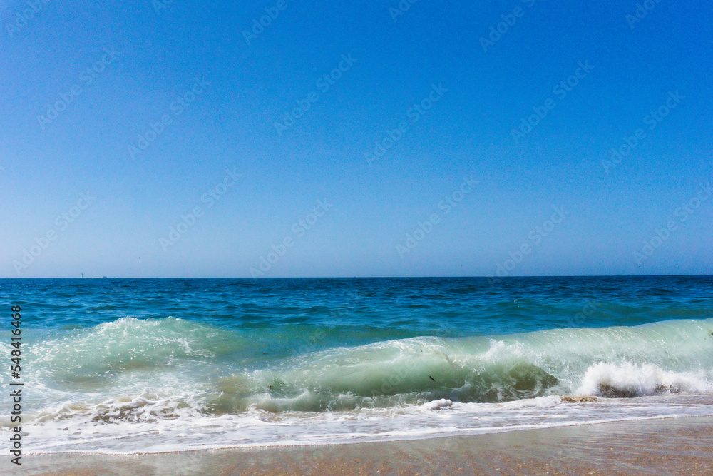 Waves breaking onto sandy beach under a blue sky