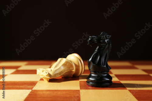 Wooden black knight and fallen one on chessboard against dark background. Competition concept