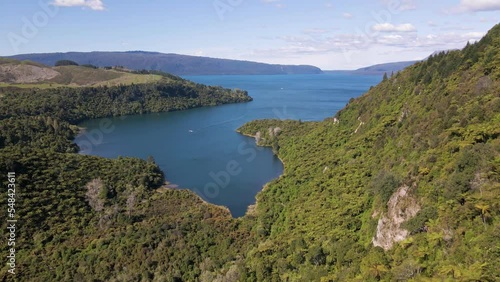 Steep and rugged cliffs that are covered by a dense palm tree forest falling off to the stunning blue lake Tarawera, New Zealand. Aerial establishing shot photo