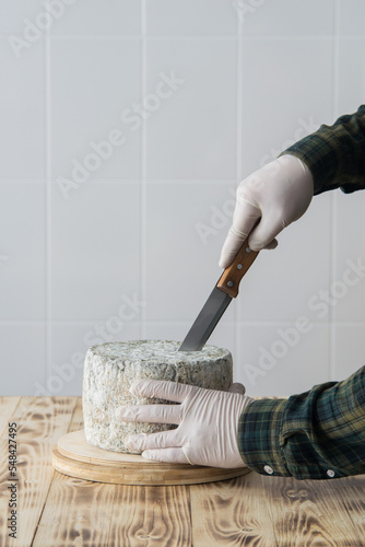 Male hands cutting round blue cheese wheel on wooden cutting board on light background, selective focus