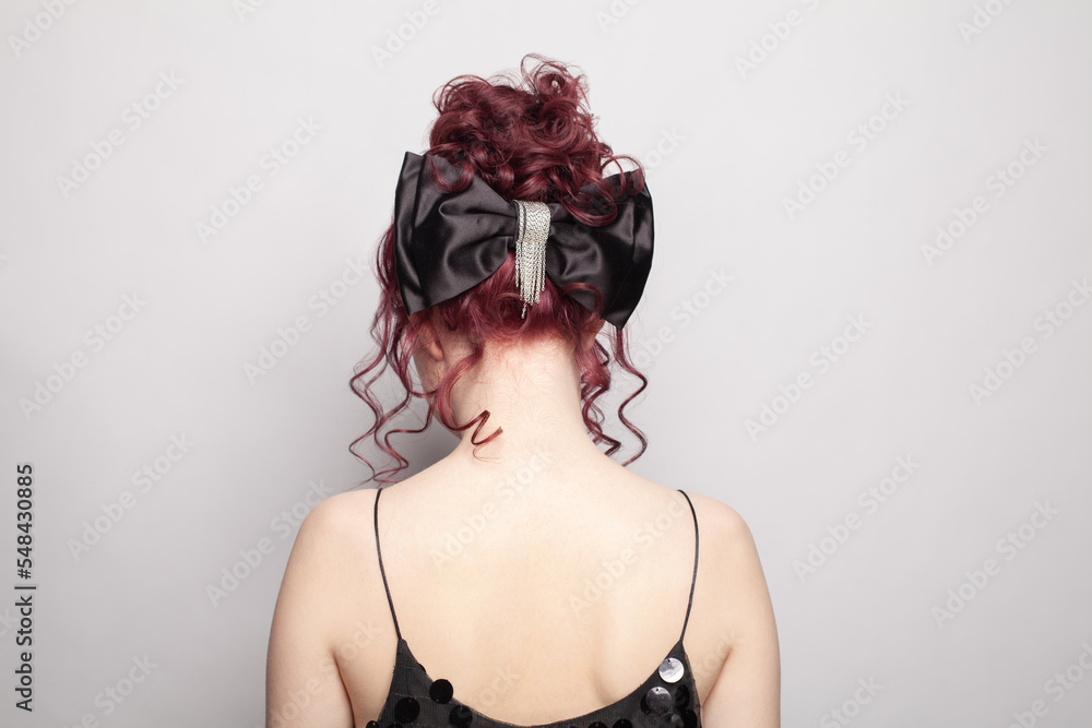 Portrait of beautiful woman with red curly hair in black dress standing with her back  against grey background