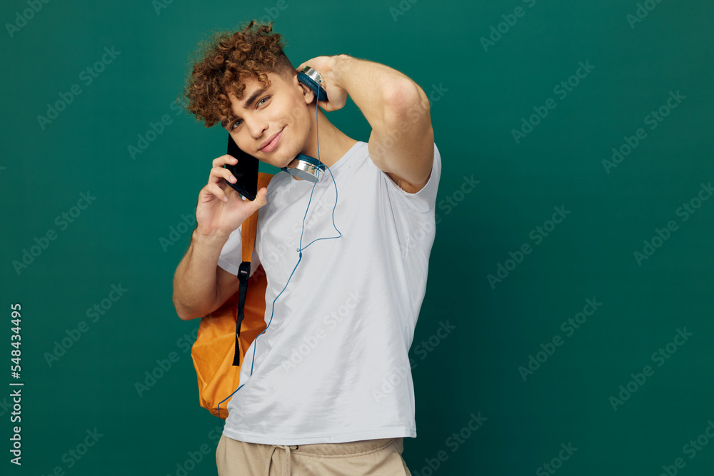 horizontal portrait of a handsome man with curly hair standing on a green background in a gray T-shirt, holding headphones around his neck, talking enthusiastically on the phone horizontal portrait of a handsome man with curly hair standing on a green background in a gray T-shirt, holding headphones around his neck, talking enthusiastically on the phone