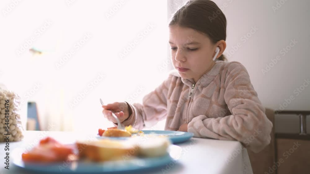 Little pretty girl in headphones has breakfast at home at the table in the kitchen. Domestic, lifestyle Little pretty girl in headphones has breakfast at home at the table in the kitchen. Domestic, lifestyle