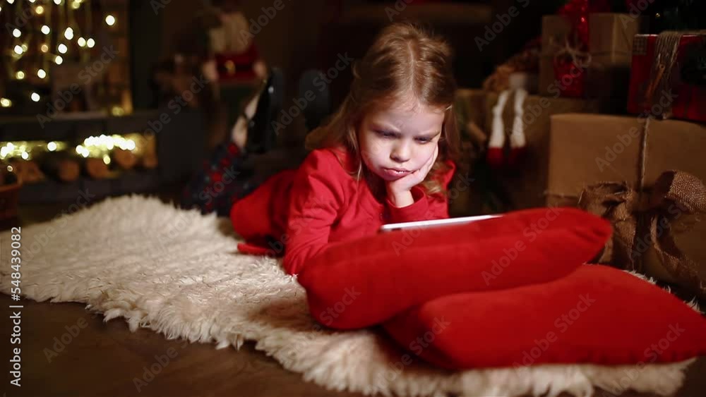 Little girl lying on the carpet near Christmas gifts, concentrated using tablet. Camera moving Little girl lying on the carpet near Christmas gifts, concentrated using tablet. Camera moving