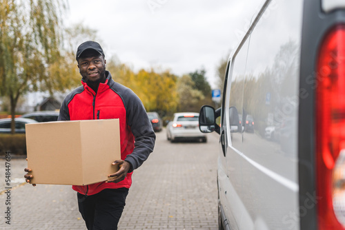 Young adult black delivery guy wearing red uniform pullover walking to white van carrying cardboard box package. Horizontal outdoor shot. High quality photo © PoppyPix