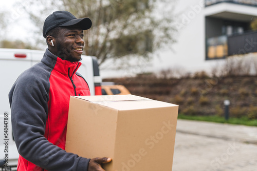 Black young adult man wearing work uniform red pullover smiling carrying cardboard box package to deliver. Horizontal outdoor shot . High quality photo
