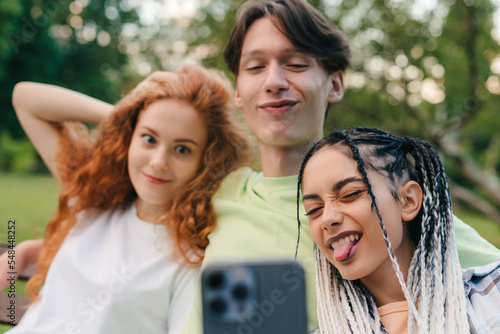 Cheerful smiling teenagers relaxing at the park on sunny summer day and taking selfies with mobile phone. Modern distance communication concept. Three people.