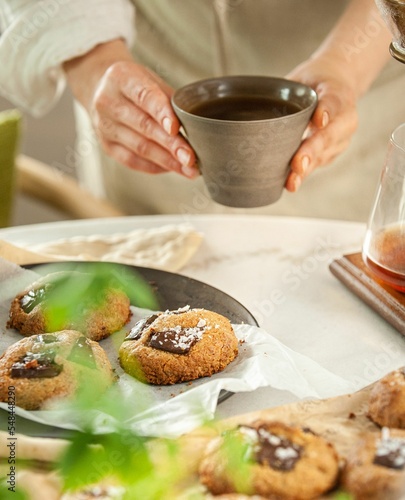 Vertical closeup of female hands holding coffee cup and freshly baked cookies blurred background