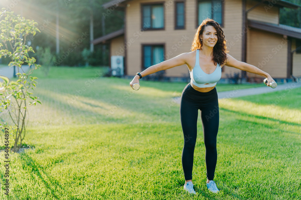 Slim motivated brunette woman dressed in cropped top and leggings, has workout with dumbbells, poses on green lawn near private house, has perfect body shape. Healthy lifestyle and sport concept