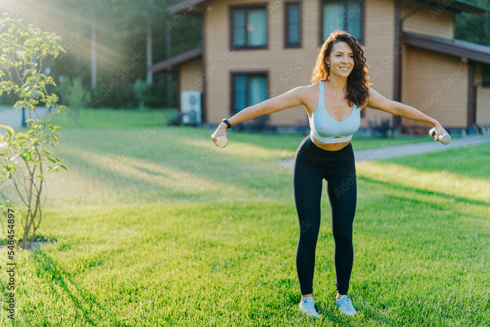 Slim motivated brunette woman dressed in cropped top and leggings, has workout with dumbbells, poses on green lawn near private house, has perfect body shape. Healthy lifestyle and sport concept Slim motivated brunette woman dressed in cropped top and leggings, has workout with dumbbells, poses on green lawn near private house, has perfect body shape. Healthy lifestyle and sport concept