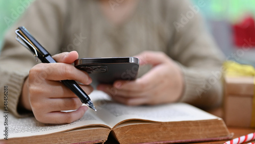 A woman in cozy sweater using her smartphone to search online information while reading a book.