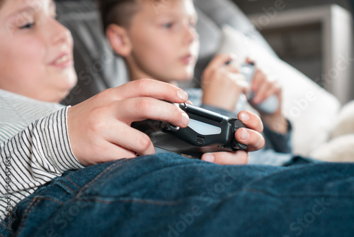 Side view of two teenage boys lying on sofa at home, holding gaming controller joystick gamepad, playing videogames on console station. Hobby, free time, gaming, entertainment, friendship. Soft focus.