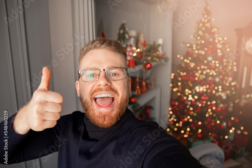 Happy lonely young man in glasses takes selfie photo on background Christmas interior house