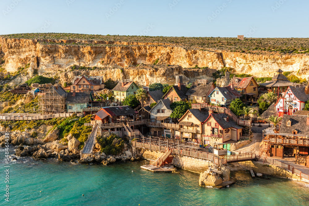 Malta, Il-Mellieha. View of the famous Popeye Village in Anchor Bay. Turquoise sea waters, blue sky and sunset light on cliffs