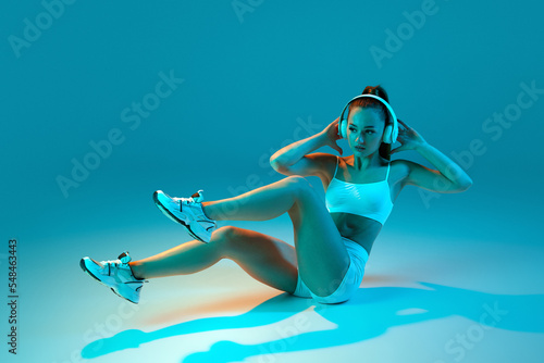 Young sportive girl in white sportswear and headphones training, doing crunches isolated over blue studio background in neon light