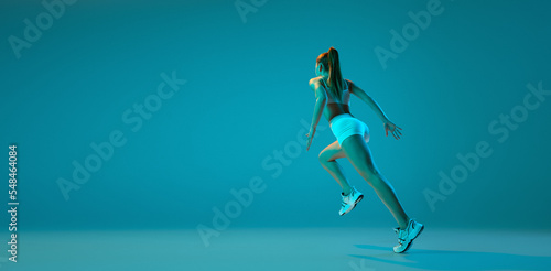 Young sportive girl in white sportswear training  running isolated over blue studio background in neon light. Flyer