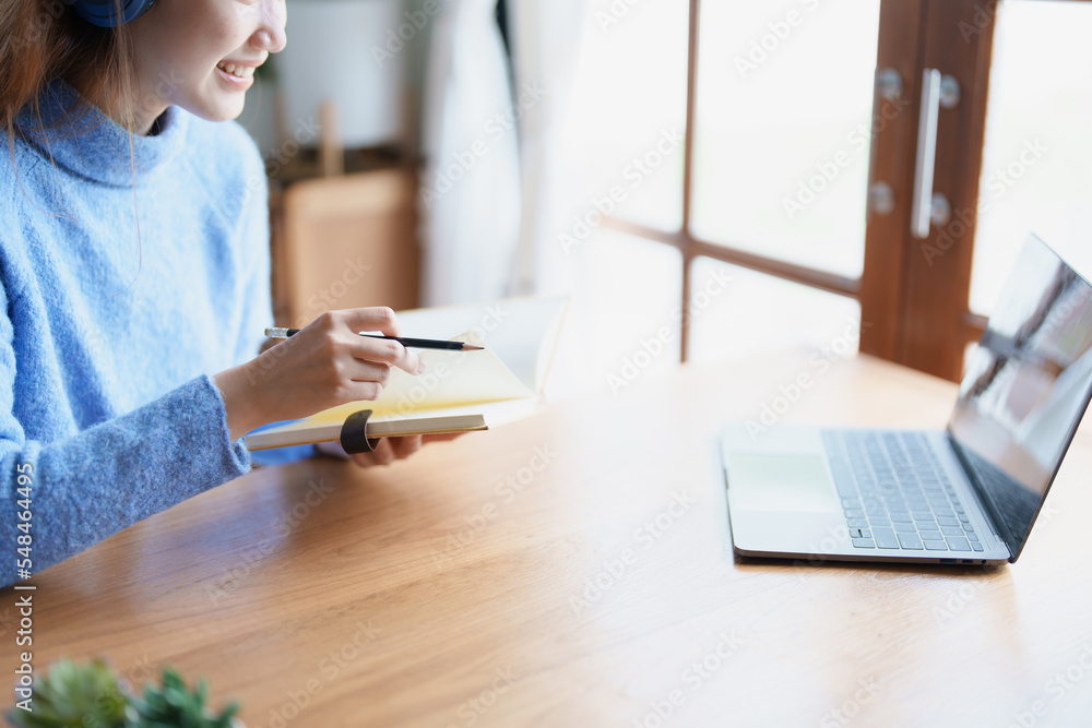 Portrait of a teenage Asian woman using a computer, wearing headphones and using a notebook to study online via video conferencing on a wooden desk at home Portrait of a teenage Asian woman using a computer, wearing headphones and using a notebook to study online via video conferencing on a wooden desk at home