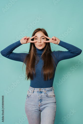 Vertical shoot of caucasian woman with long loose hair in blue sweater and blue jeans posing at studio over turquoise backdrop making victory sign near eyes by both hands. Mockup beauty and fashion.