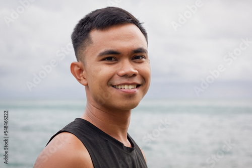 Smiling portrait of young Filipino man by the sea on cloudy day. Asian male model concept