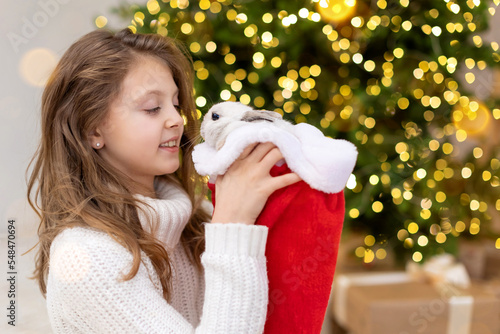 Cute Christmas bunny Symbol of the New year 2022 close-up in Santa hat in the hands of happy child on the background of a decorated Christmas tree with defocus lights