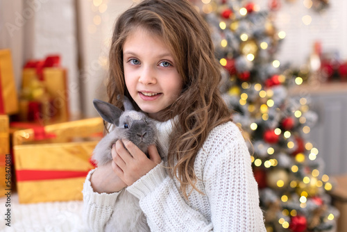 Happy Christmas bunny Symbol of the New year in the hands of a little girl on the background of a decorated Christmas tree with defocus lights