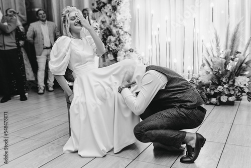 Εκτύπωση καμβά Stylish groom in brown suit pulling out garter poking his head under dress to smiling, laughing, happy blonde bride indoors