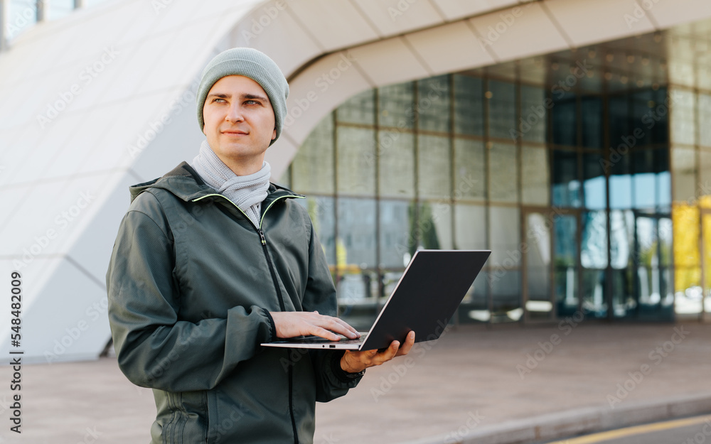 Handsome european young male using laptop while working outdoors. Student or freelancer in outerwear standing in front of university or business office and looking away Handsome european young male using laptop while working outdoors. Student or freelancer in outerwear standing in front of university or business office and looking away