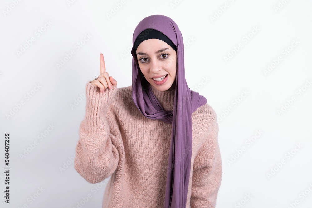 beautiful muslim woman wearing hijab and warm jumper over white background pointing finger up and looking inspired by genius thought, showing good idea sign, having clever solution in mind beautiful muslim woman wearing hijab and warm jumper over white background pointing finger up and looking inspired by genius thought, showing good idea sign, having clever solution in mind