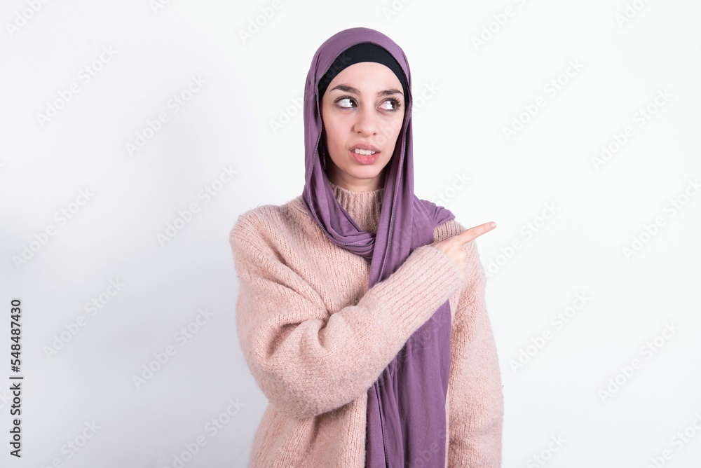 beautiful muslim woman wearing hijab and warm jumper over white background Pointing aside worried and nervous with forefinger, concern and surprise concept. beautiful muslim woman wearing hijab and warm jumper over white background Pointing aside worried and nervous with forefinger, concern and surprise concept.