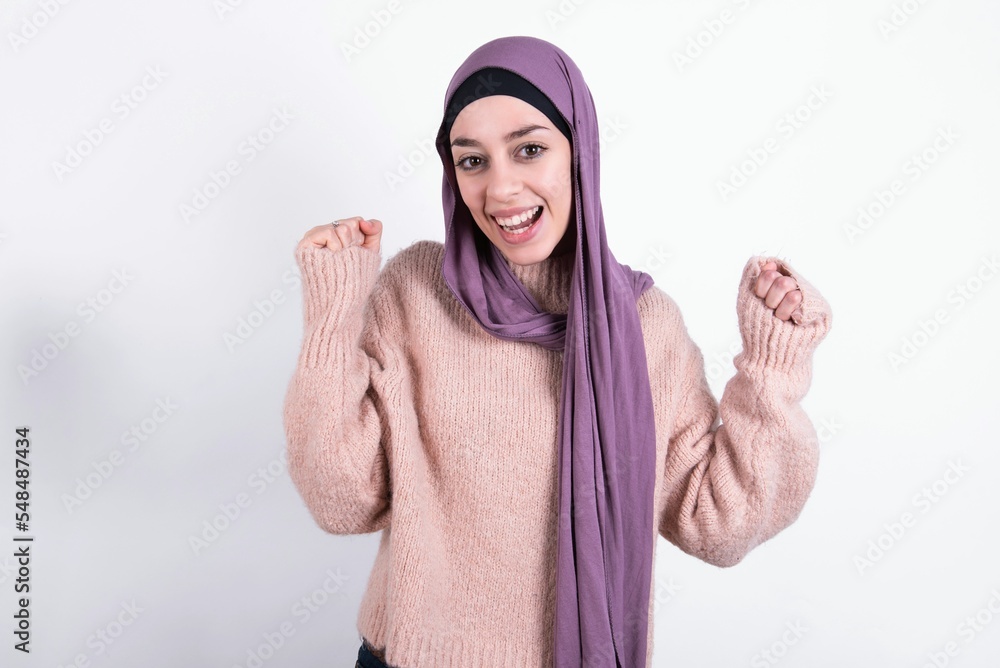 beautiful muslim woman wearing hijab and warm jumper over white background celebrating surprised and amazed for success with arms raised and open eyes. Winner concept. beautiful muslim woman wearing hijab and warm jumper over white background celebrating surprised and amazed for success with arms raised and open eyes. Winner concept.