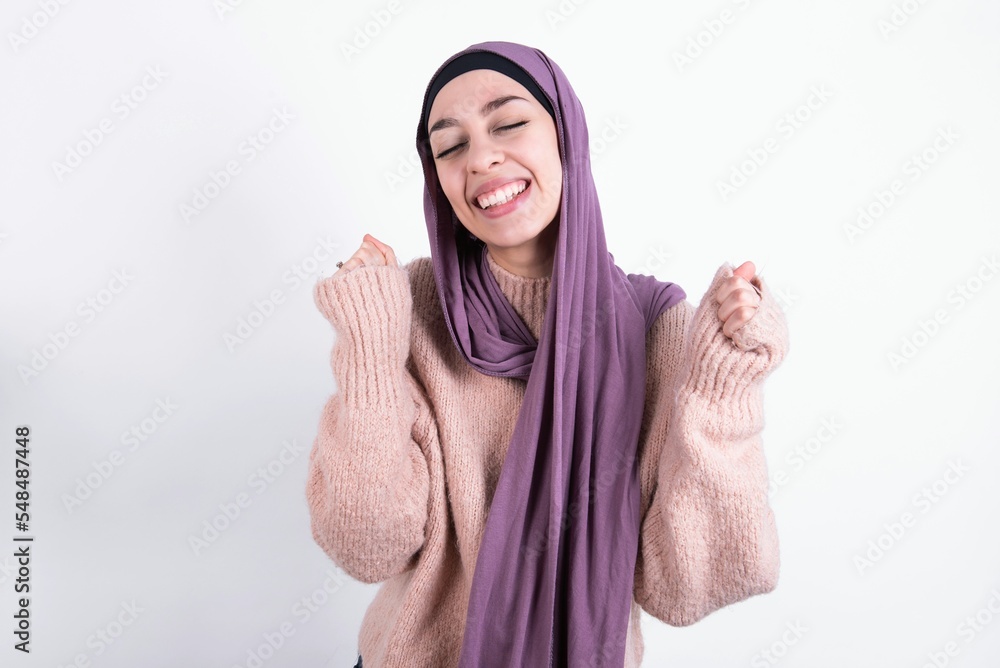 Φωτογραφία beautiful muslim woman wearing hijab and warm jumper over white background being excited for success with raised arms and closed eyes celebrating victory