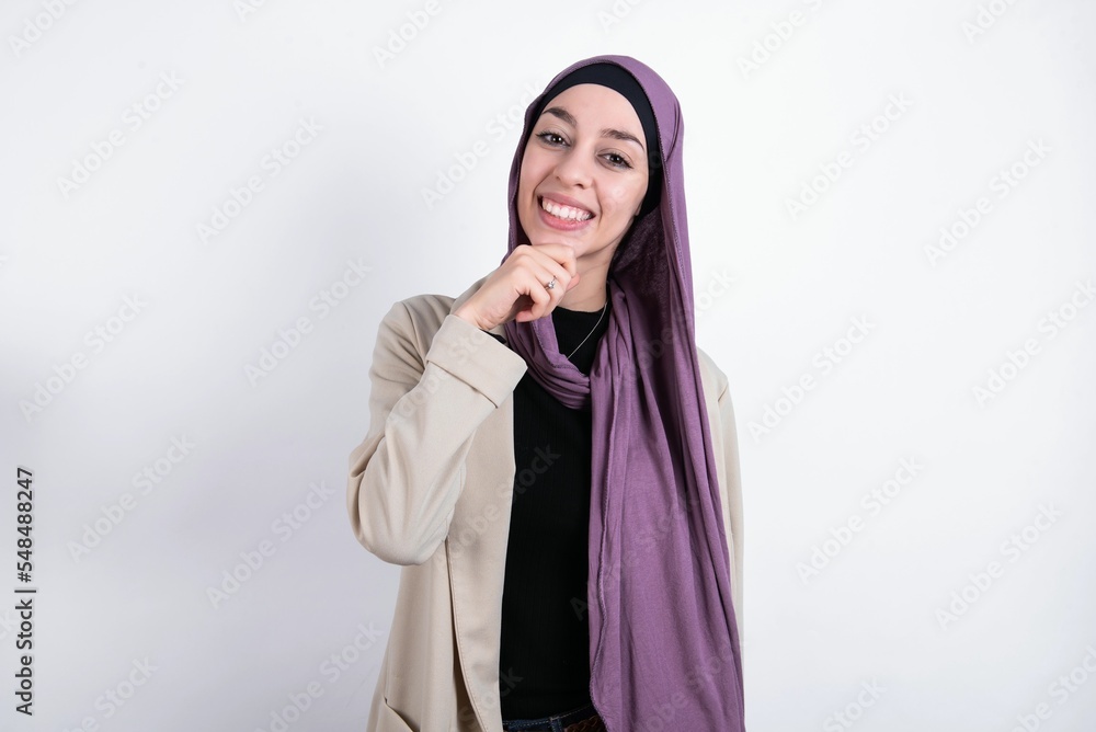 young beautiful muslim woman wearing hijab and jacket over white background laughs happily keeps hand on chin expresses positive emotions smiles broadly has carefree expression young beautiful muslim woman wearing hijab and jacket over white background laughs happily keeps hand on chin expresses positive emotions smiles broadly has carefree expression