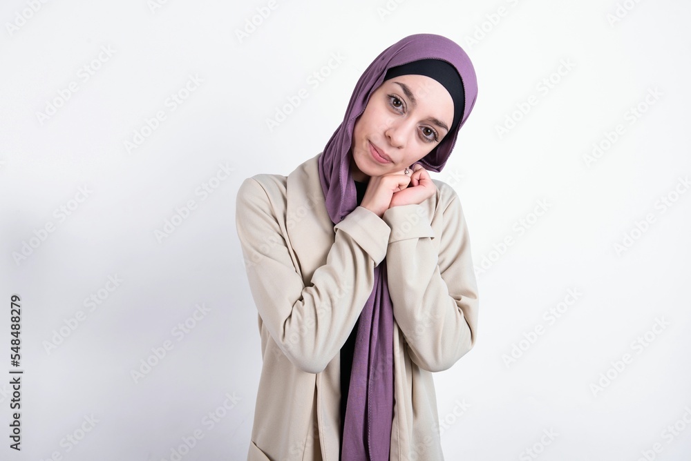 Charming serious young beautiful muslim woman wearing hijab and jacket over white background keeps hands near face smiles tenderly at camera Charming serious young beautiful muslim woman wearing hijab and jacket over white background keeps hands near face smiles tenderly at camera