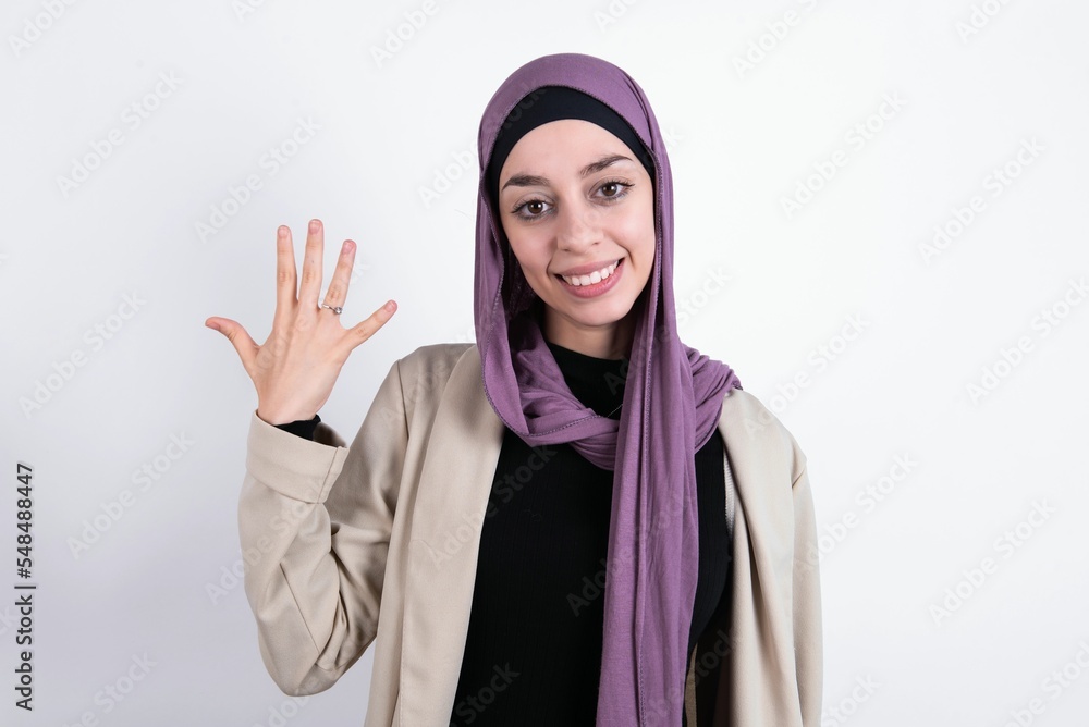 young beautiful muslim woman wearing hijab and jacket over white background smiling and looking friendly, showing number five or fifth with hand forward, counting down young beautiful muslim woman wearing hijab and jacket over white background smiling and looking friendly, showing number five or fifth with hand forward, counting down