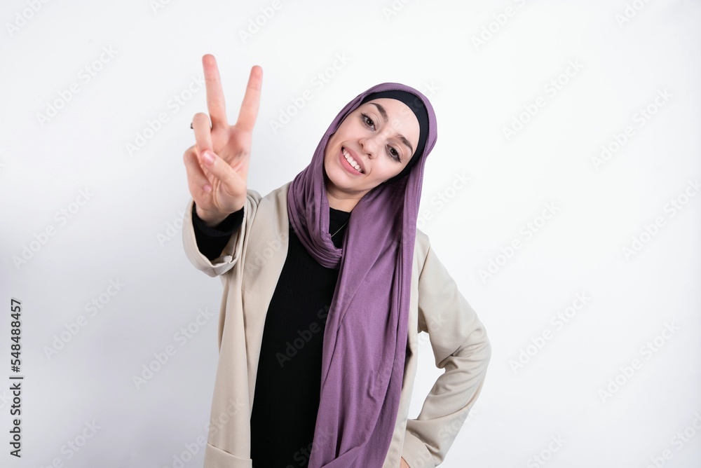 young beautiful muslim woman wearing hijab and jacket over white background smiling and looking happy, carefree and positive, gesturing victory or peace with one hand young beautiful muslim woman wearing hijab and jacket over white background smiling and looking happy, carefree and positive, gesturing victory or peace with one hand