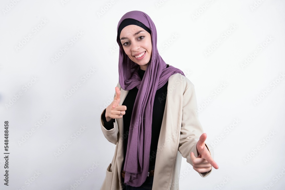 young beautiful muslim woman wearing hijab and jacket over white background directs fingers at camera selects someone. I recommend you. Best choice young beautiful muslim woman wearing hijab and jacket over white background directs fingers at camera selects someone. I recommend you. Best choice