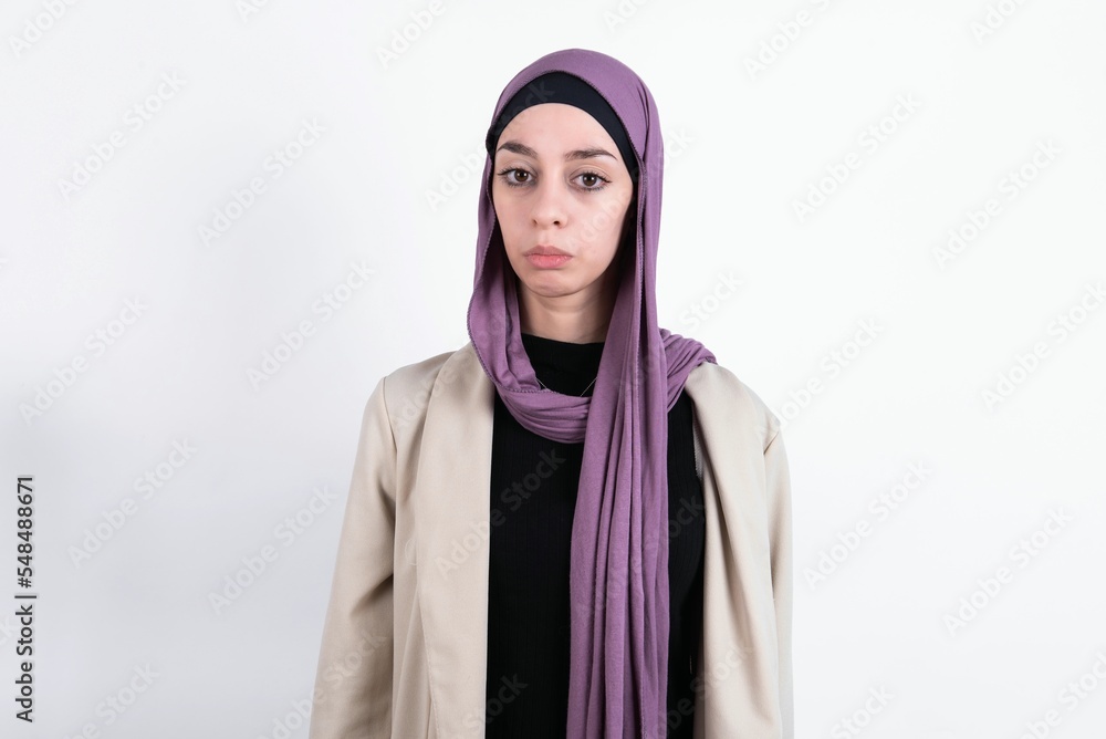 Dissatisfied young beautiful muslim woman wearing hijab and jacket over white background purses lips and has unhappy expression looks away stands offended. Depressed frustrated model. Dissatisfied young beautiful muslim woman wearing hijab and jacket over white background purses lips and has unhappy expression looks away stands offended. Depressed frustrated model.