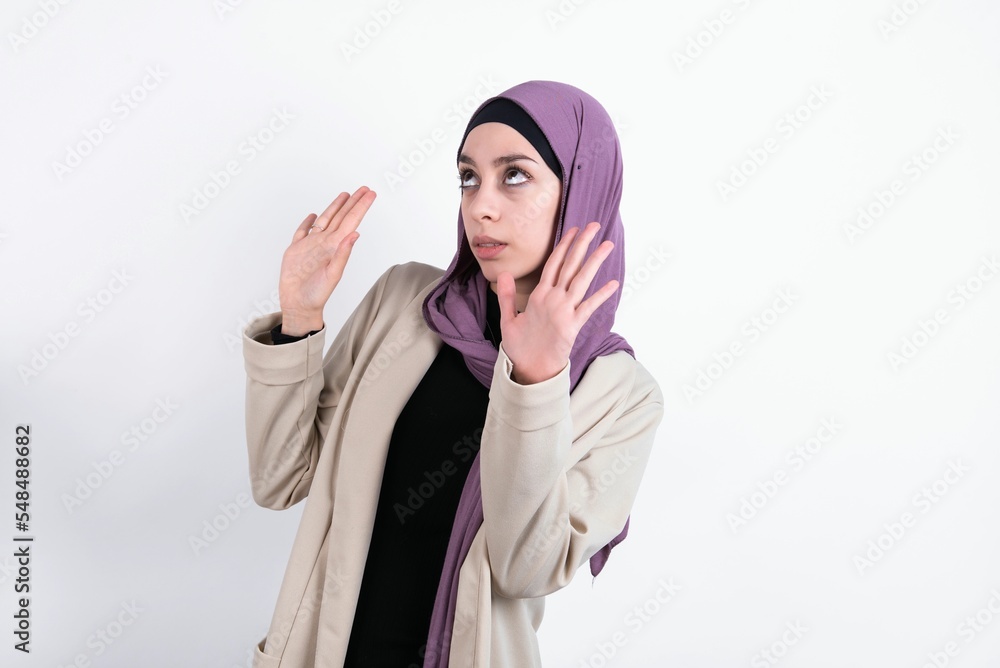 young beautiful muslim woman wearing hijab and jacket over white background keeps palms forward and looks with fright above on ceiling tries to defense herself from invisible danger opens mouth. young beautiful muslim woman wearing hijab and jacket over white background keeps palms forward and looks with fright above on ceiling tries to defense herself from invisible danger opens mouth.
