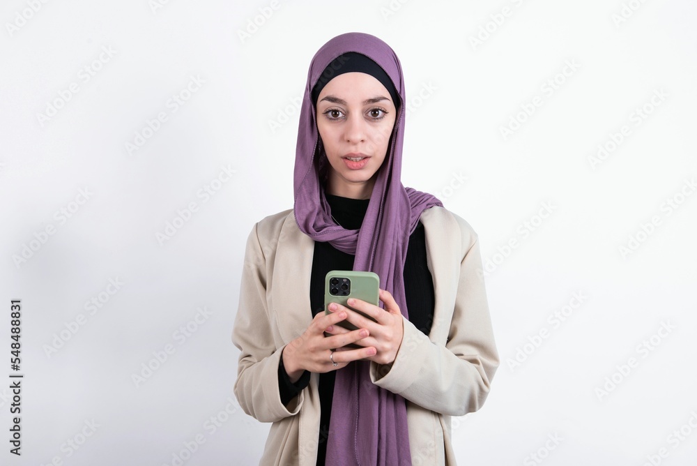young beautiful muslim woman wearing hijab and jacket over white background holds mobile phone in hands and rejoices positive news, uses modern cellular young beautiful muslim woman wearing hijab and jacket over white background holds mobile phone in hands and rejoices positive news, uses modern cellular