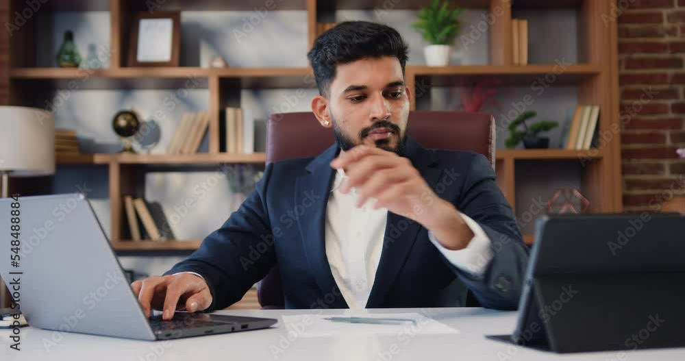 Handsome multitasking Indian businessman in formal suit working using the touchpad while typing on a laptop and taking notes on computer tablet at modern office Handsome multitasking Indian businessman in formal suit working using the touchpad while typing on a laptop and taking notes on computer tablet at modern office