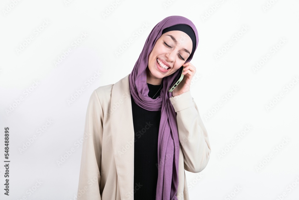 Φωτογραφία Funny young beautiful muslim woman wearing hijab and jacket over white background laughs happily, has phone conversation, being amused by friend, closes eyes