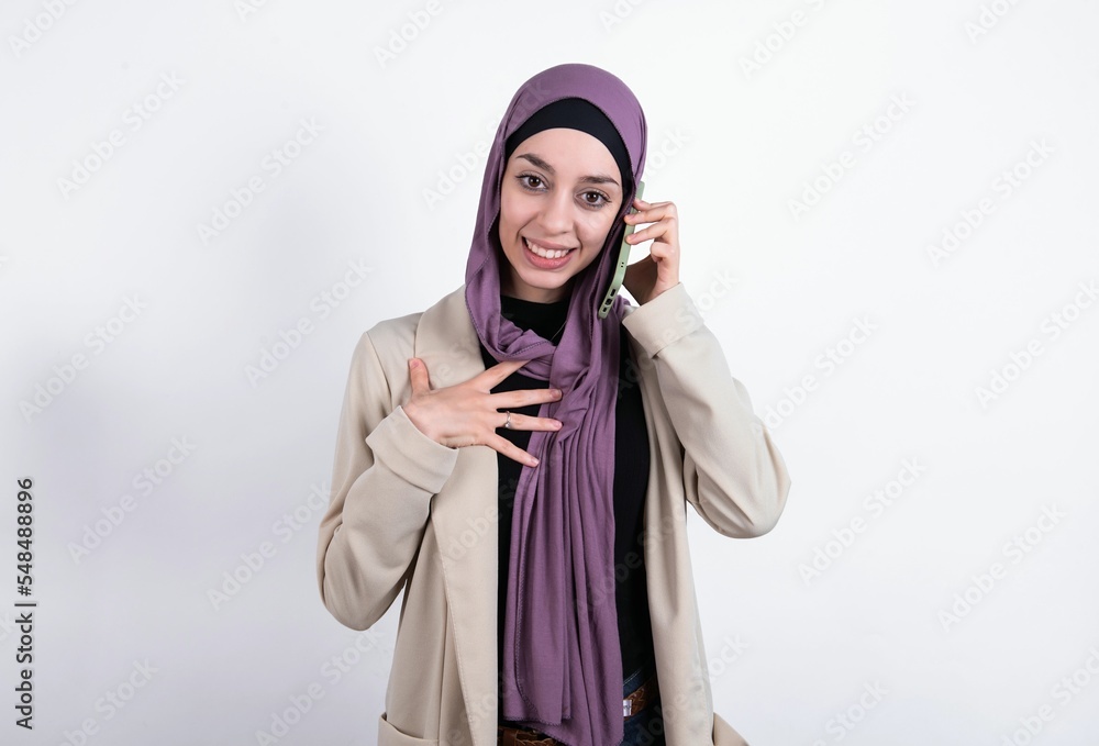 Φωτογραφία Smiling young beautiful muslim woman wearing hijab and jacket over white background talks via cellphone, enjoys pleasant great conversation