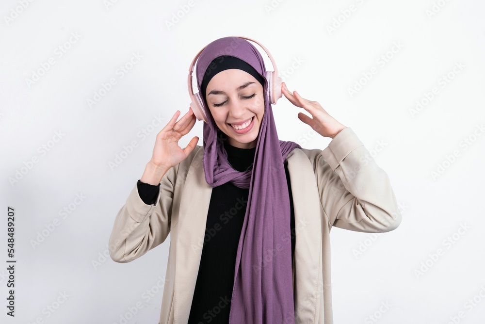 Pleased young beautiful muslim woman wearing hijab and jacket over white background enjoys listening pleasant melody keeps hands on stereo headphones closes eyes. Spending free time with music Pleased young beautiful muslim woman wearing hijab and jacket over white background enjoys listening pleasant melody keeps hands on stereo headphones closes eyes. Spending free time with music
