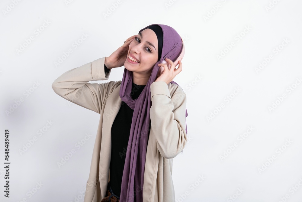 young beautiful muslim woman wearing hijab and jacket over white background wears stereo headphones listens music concentrated aside. People hobby lifestyle concept young beautiful muslim woman wearing hijab and jacket over white background wears stereo headphones listens music concentrated aside. People hobby lifestyle concept