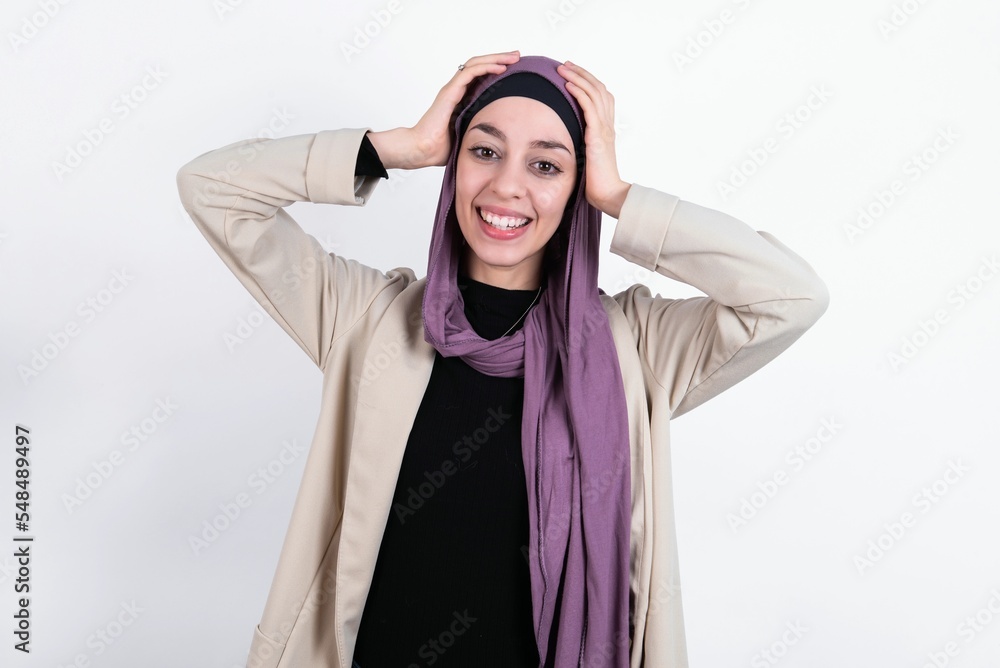 Cheerful overjoyed young beautiful muslim woman wearing hijab and jacket over white background reacts rising hands over head after receiving great news. Cheerful overjoyed young beautiful muslim woman wearing hijab and jacket over white background reacts rising hands over head after receiving great news.