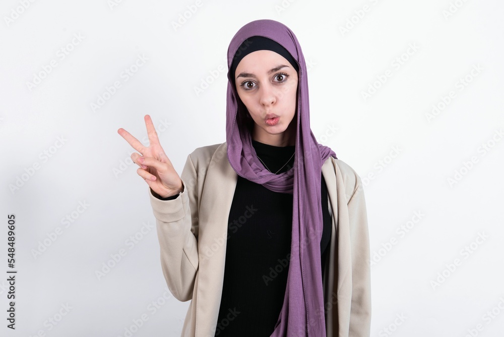 young beautiful muslim woman wearing hijab and jacket over white background makes peace gesture keeps lips folded shows v sign. Body language concept young beautiful muslim woman wearing hijab and jacket over white background makes peace gesture keeps lips folded shows v sign. Body language concept