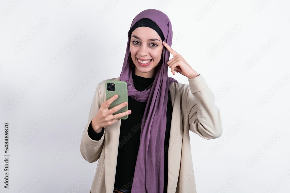 Pensive young beautiful muslim woman wearing hijab and jacket over white background keeps index finger on temple, holds modern smartphone and concentrated aside Pensive young beautiful muslim woman wearing hijab and jacket over white background keeps index finger on temple, holds modern smartphone and concentrated aside